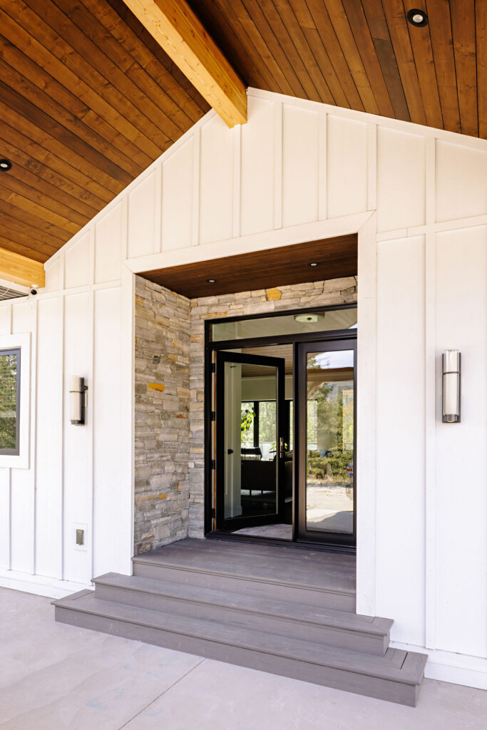Image showing entrance to a home. The siding is white board and batten style with natural stone surrounding the glass double doors.