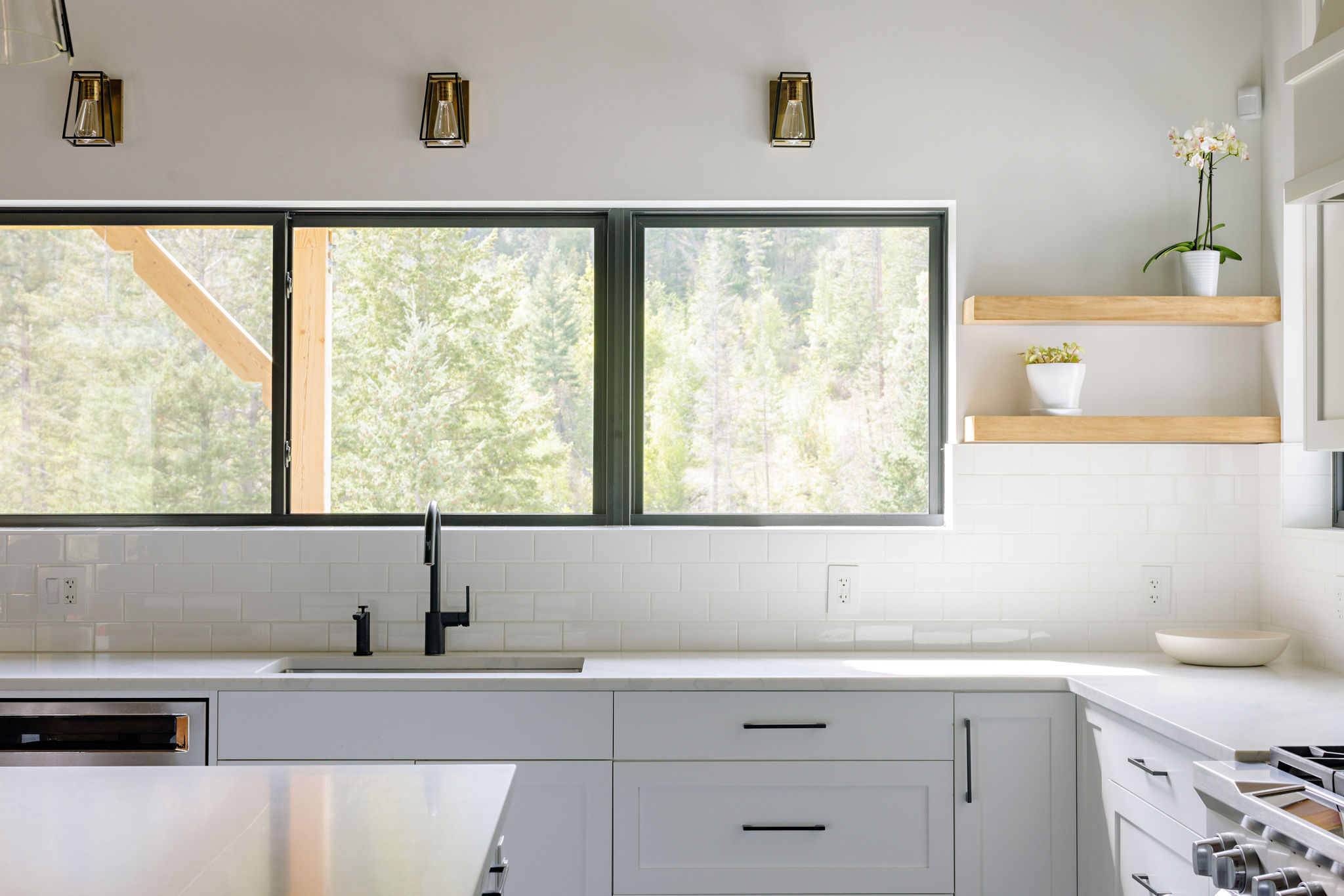 Image of the kitchen sink with white cabinetry and a row of large windows above. There are natural wood open shelves with decorative plants.
