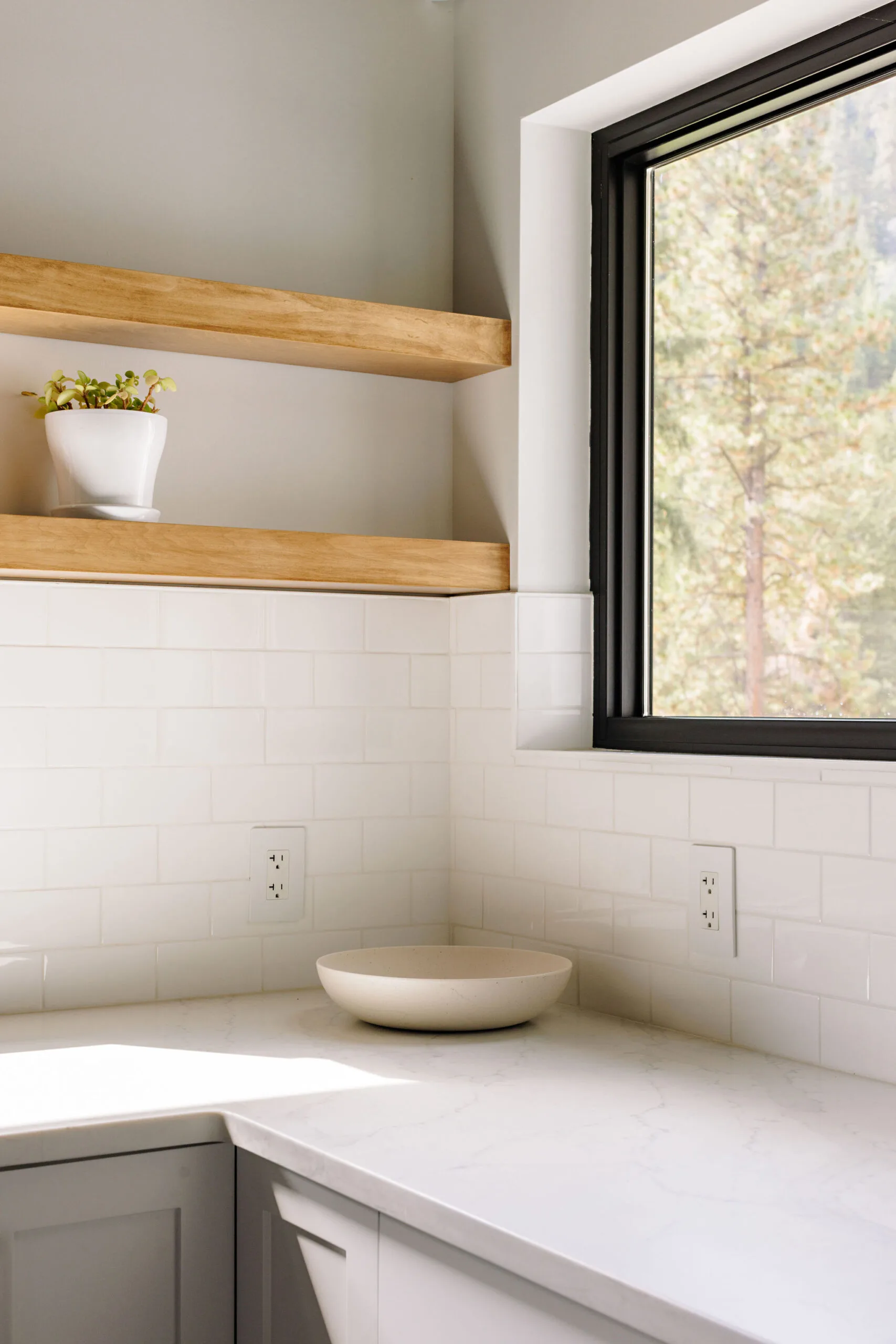 Image showing the countertops, window and open shelving in the home at Fish Lake. The counter is white stone and the backsplash is white subway tile. The open shelves are natural wood.
