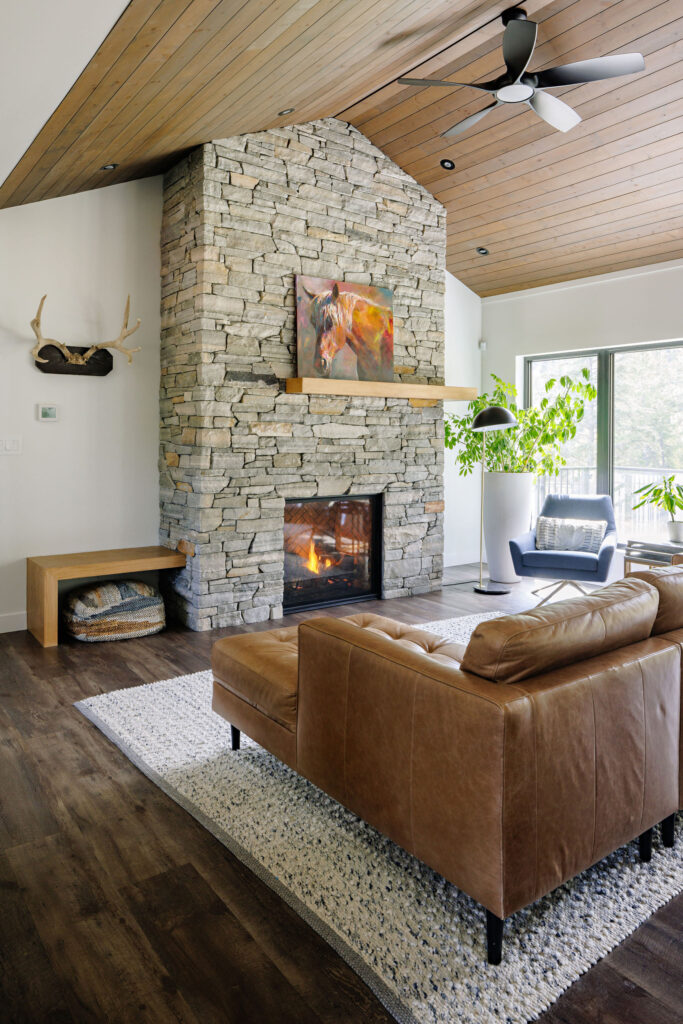 Image showing the living room with vaulted T&G wood ceiling met with a stone fireplace surround. There is a brown leather sectional and white area rug. The floor with dark brown wood.