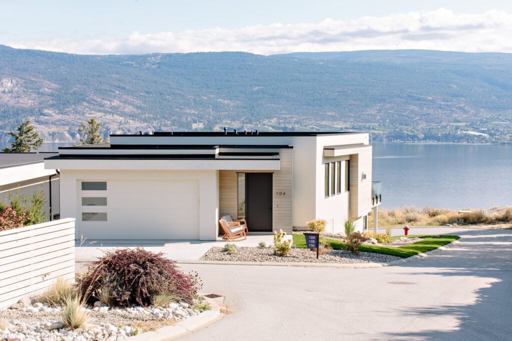 Image shoing the exxterior of a house built into the hillside with lake and mountainer views. The house is white with a flat black roof. Built by Whitfield Homes