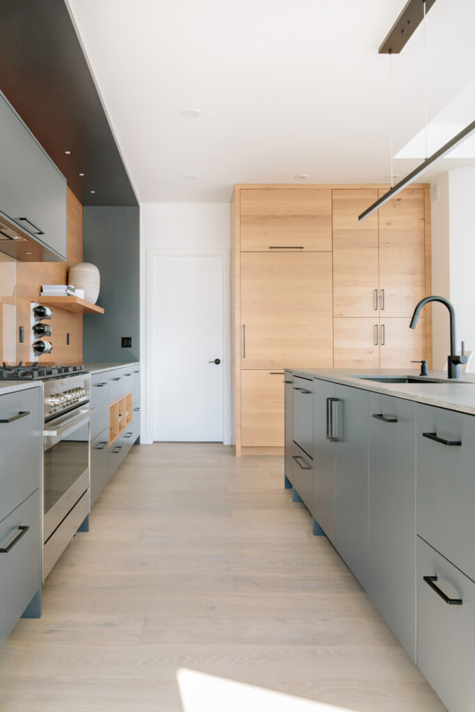 Image showing custom grey cabinetry with a Caesarstone countertop. There is a stainless steel natural gas stove and a natural wood backsplash. The island has a black sink and faucet and there is a bannk of full height cabinets along the far wall that are natural wood. Built by Whitfield Homes