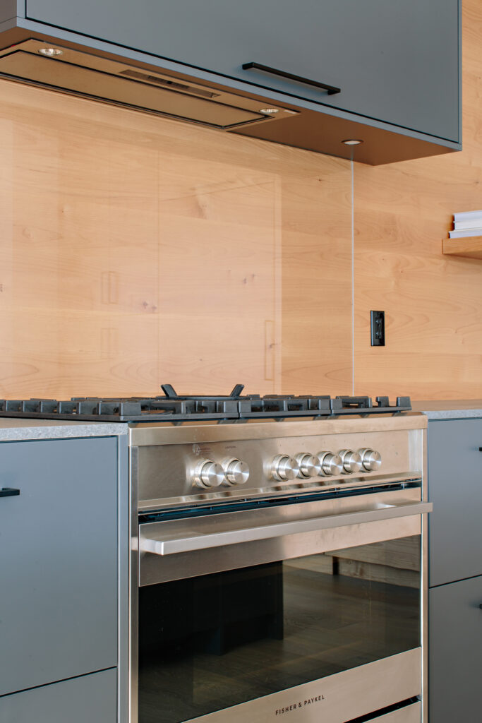 Image showing the chef style stainless steel stove in the kitchen. There are grey cabinets and a natural wood backsplash. Built by Whitfield Homes.