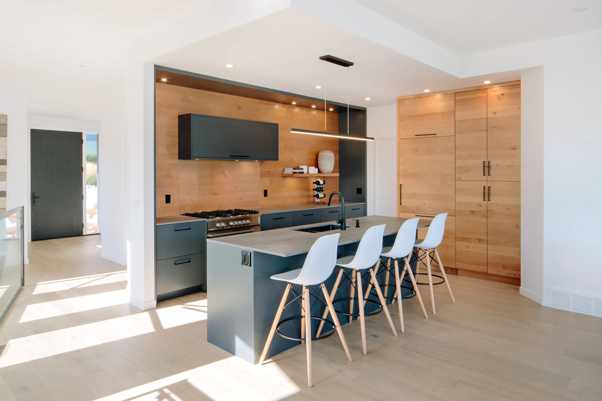 Image of a kitchen featuring custom grey cabinets with a wood grain backsplash and Caesarstone countertops. The island has a sink and breakfast bar with four white stools. Built by Whitfield Homes