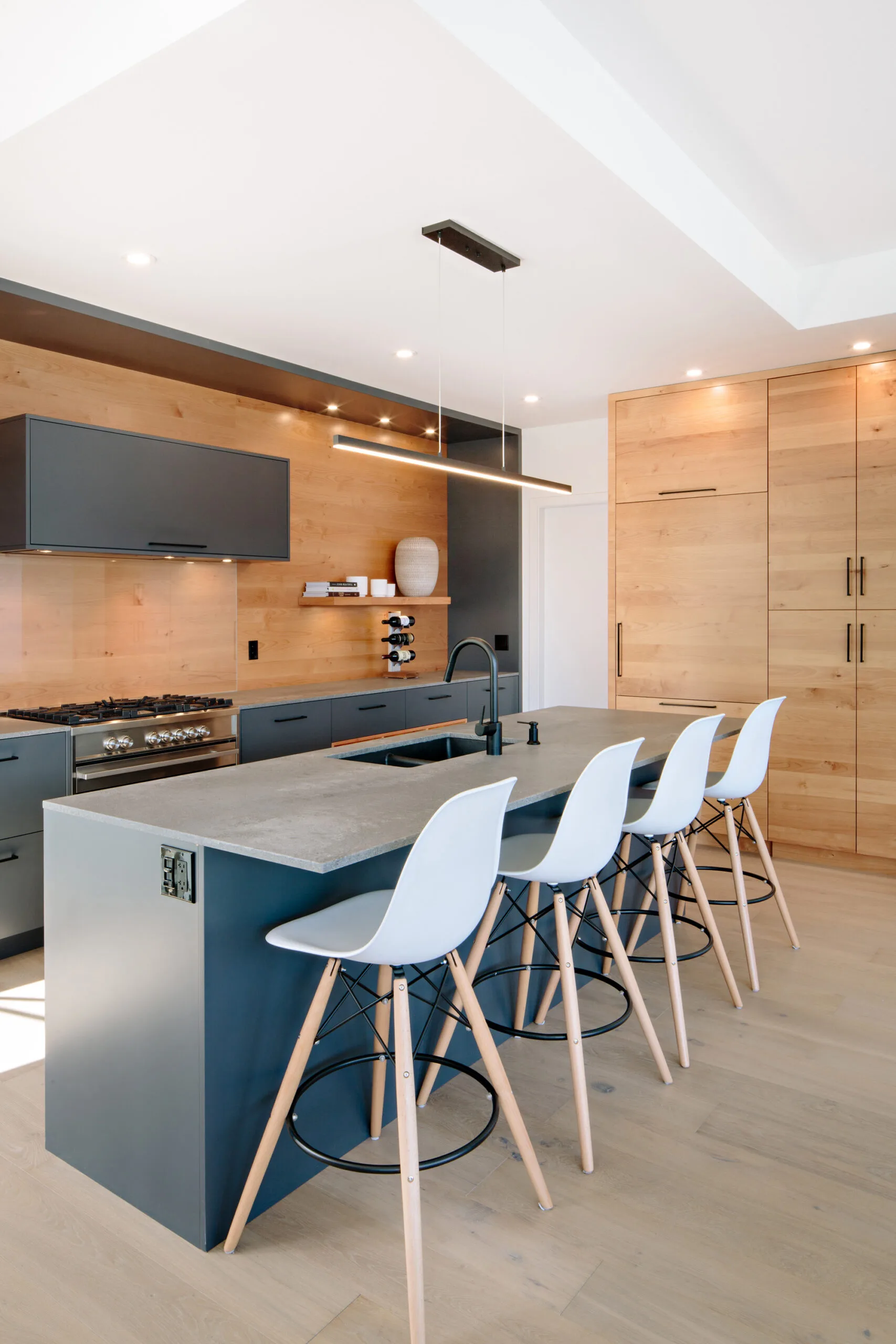 Image of a kitchen featuring custom grey cabinets with a wood grain backsplash and Caesarstone countertops. The island has a sink and breakfast bar with four white stools. Built by Whitfield Homes