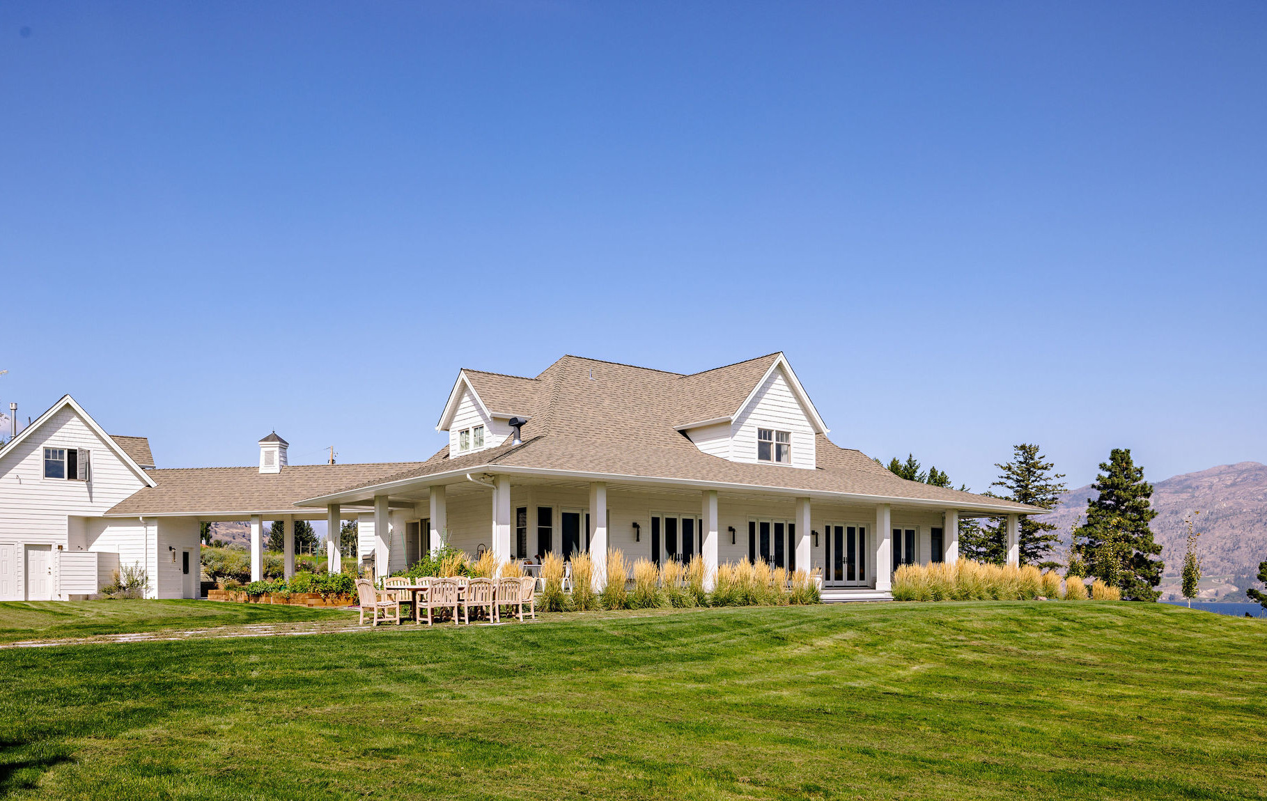 Image of the exterior of a white farmhouse with a wraparound porch and a separated garage connected by a covered walkway. Renovated by Whitfield Homes.