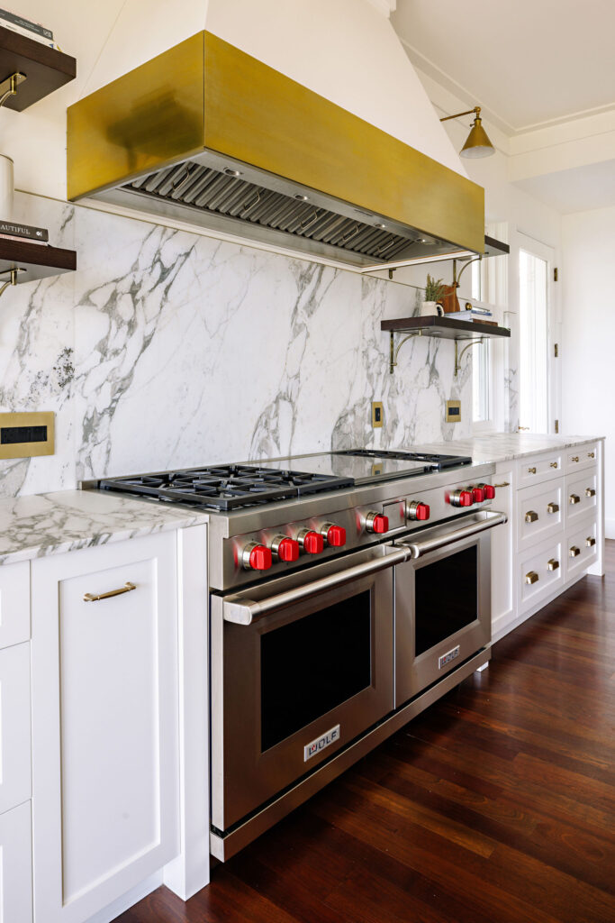 Image showing the cooking area with a large six burner range, vent hood with brass details and open shelving with display items.